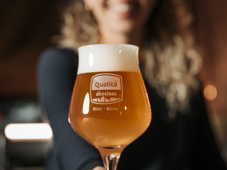 © IDM Südtirol/Daniele Fiorentino Close-up of a beer glass engraved with the South Tyrol seal of quality, a girl holding the glass in the background. | © IDM Südtirol/Daniele Fiorentino