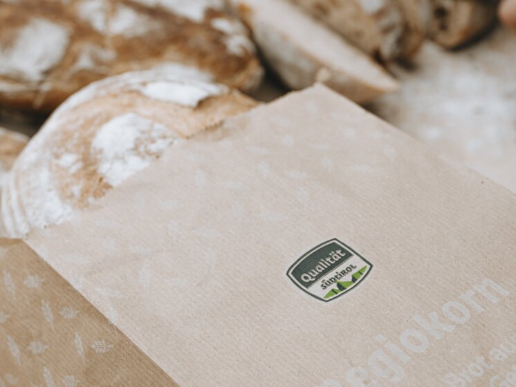 © IDM Südtirol/Daniele Fiorentino A bread bag with the South Tyrol seal of quality,in the background various types of bread and a person slicing bread. | © IDM Südtirol/Daniele Fiorentino